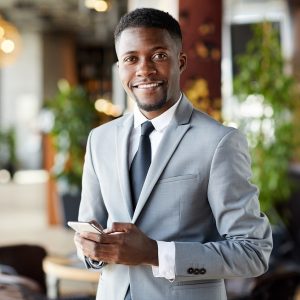 Portrait of cheerful confident handsome young African-American businessman with beard using smartphone to text message